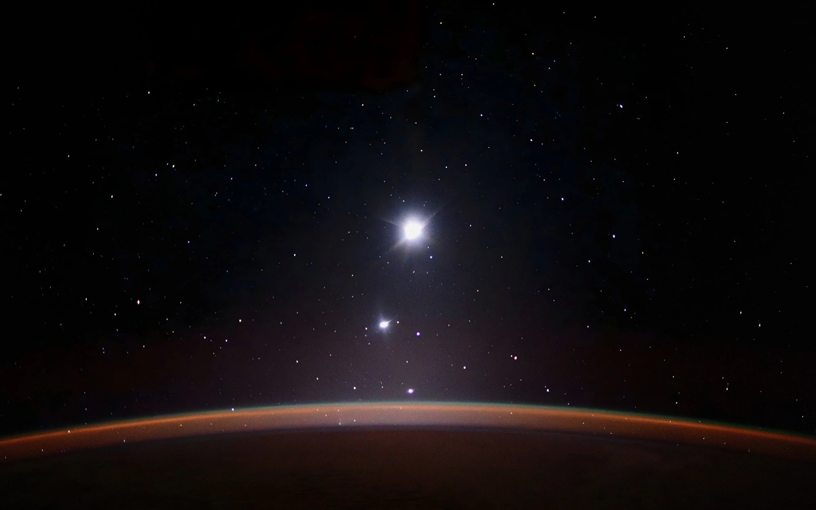 Earth's limb glowing orange at night under a dark sky with a few bright stars and a bright planet.