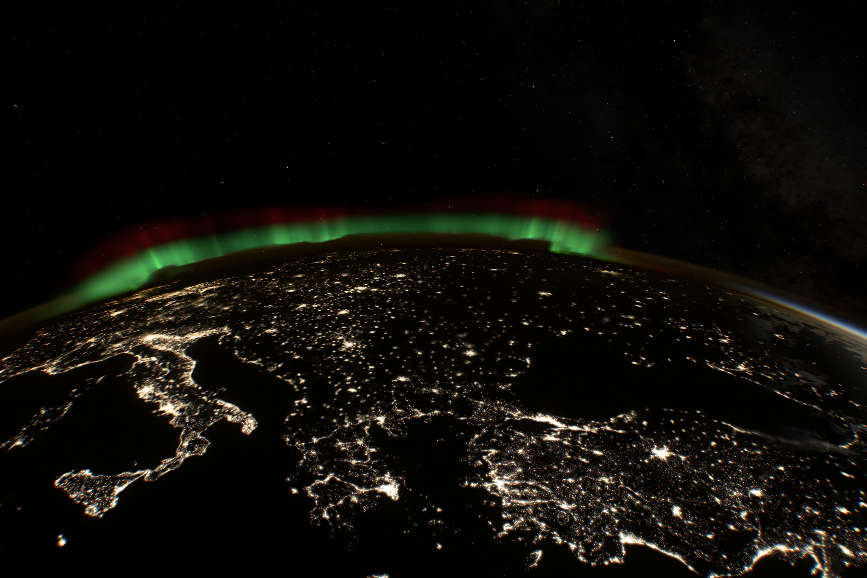 Europe at night from orbit: Italy, the Balkans and the cities of central Europe as clusters of light joined by fainter roads, with a green and red aurora along the far horizon.