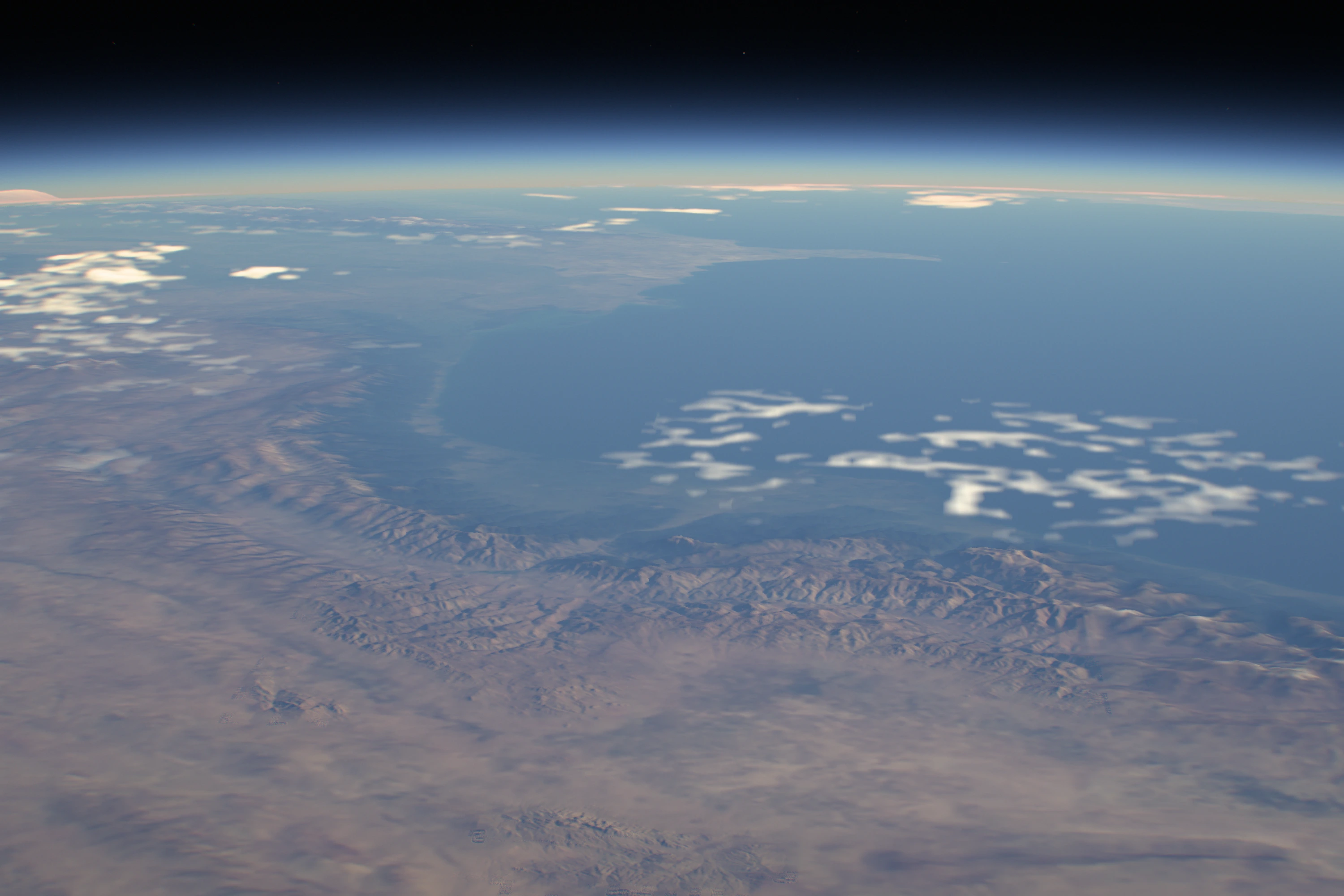 A morning coastline at 2.25×: a sea under a blue limb, ridged mountains in low sunlight and small bright clouds over the water.