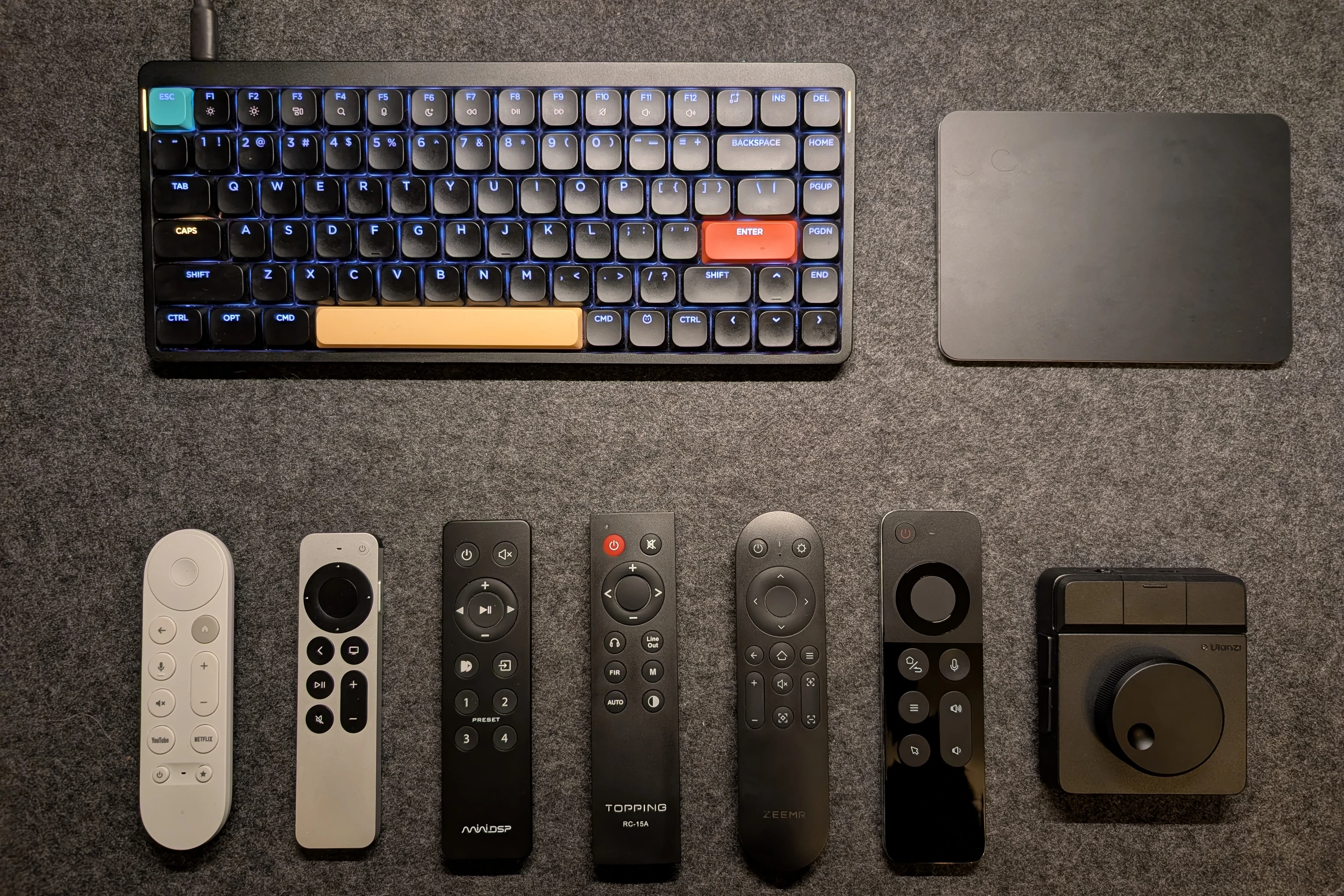 Seen from above on a grey felt mat: a NuPhy keyboard with a blue backlight and a black Magic Trackpad, and below them a row of seven remotes, a white Google TV remote, the silver Siri Remote, black remotes for the miniDSP, a Topping DAC and a Zeemr projector, the glossy Air Mouse and the square Ulanzi Dial.